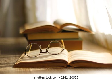 Open book and reading glasses on the table, illuminated by sunlight. Stack of vintage books in the background. Selective focus. - Powered by Shutterstock