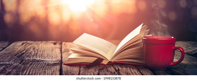 Open Book And Steaming Cup Of Coffee On Wooden Table With Autumn Sunrise Background - Study And Meditation Concept - Powered by Shutterstock