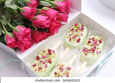 Open Box With Cakes In The Form Of Popsicle On A Stick In Light Green Glaze And Flower And Pistachio Topping. And A Large Bouquet Of Bright Pink Roses.
