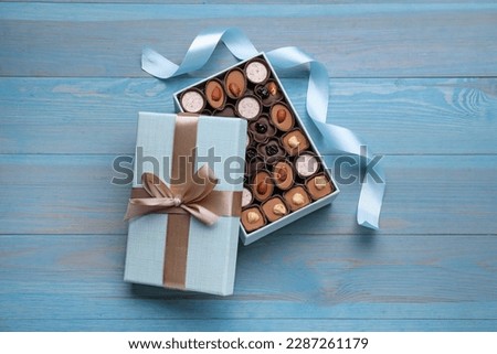 Open box of delicious chocolate candies and color ribbon on light blue wooden table, flat lay