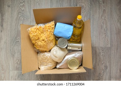 Open Box Of Grocery Stands On The Wooden Floor In The Center: Oil, Rice, Pasta, Canned Food, Cornflakes. Food Help  To Schoolchildren. Top View. Horizontal Oriented. 