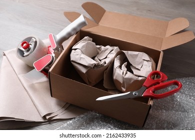 Open Box With Wrapped Items, Adhesive Tape, Scissors, Paper And Bubble Wrap On Wooden Table