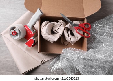 Open box with wrapped items, adhesive tape, scissors, paper and bubble wrap on wooden table, flat lay - Shutterstock ID 2163543323