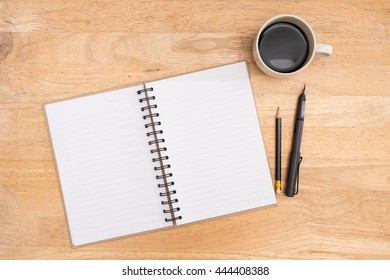 open cap matte black calligraphic pen beside recycle paper notebook black sharpened wood pencil and hot black coffee on light brown jointed rubberwood desktop
