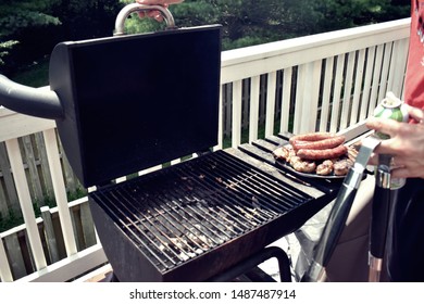 Open Charcoal Grill On A Balcony, With A Plate Of Cooked Ausage And Chicken On The Side.  With A Person Holding Tongs And Getting Ready To Close The Lid. 