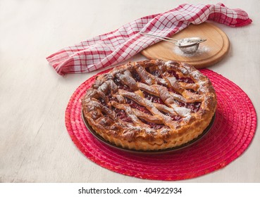 Open cherry pie on the red stand next to a sieve to sprinkle powdered sugar