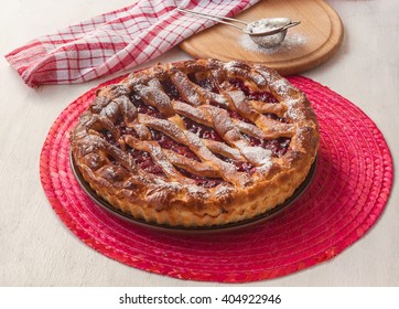 Open cherry pie on the red stand next to a sieve to sprinkle powdered sugar