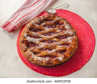 Open cherry pie on the red stand next to a sieve to sprinkle powdered sugar