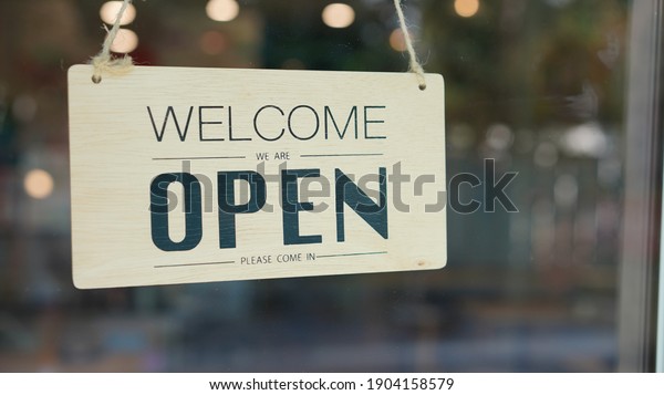 Open and closed flip sign in\
front of coffee shop and restaurant glass door. Wooden sign with\
wording of place\'s status. Say Welcome. We\'re OPEN. Please come\
in.