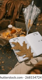 An open diary with a large leaf lies on a person's lap, with coffee and a croissant on a bench in the background, evoking a cozy autumn atmosphere.