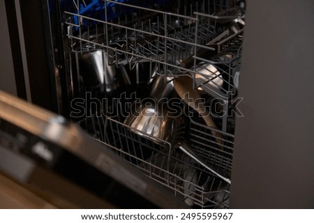 An open dishwasher packed with various kitchen items, including metal pots, utensils, and a wooden spoon. The dishwasher is in a modern kitchen setting ready for a cleaning cycle.