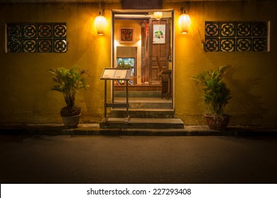 Open Door To Old Cafe In Hoi An. Restaurant At Night In Vietnam, Asia. Stand With Menu At Entrance. Plants In Pots. Light From Lamps. Traditional Asian Place To Eat. Beautiful Design Of Windows.