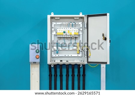 Open electrical distribution panel with circuit breakers, grounding wire, and control button box mounted on blue wall. Electrical engineering. Power control. Installation.