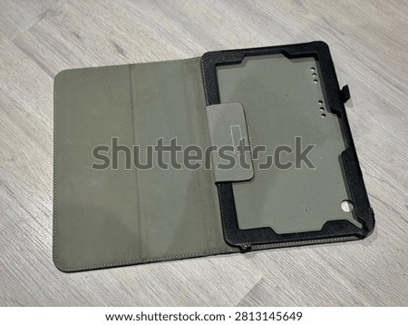 Open empty black tablet folio case showing grey suede interior lining and holder frame with corner clips on grey wood floor