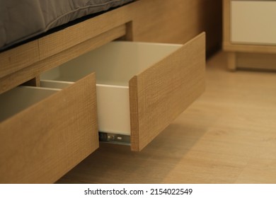 Open empty drawer. Wooden bed on laminate floor in modern bedroom. Ligth brown drawer for storage under bed. Close up bedside view for interior design. Urban life for house, real estate, living web.