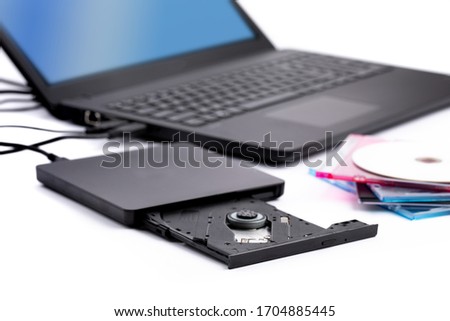 an open external computer DVD CD drive with a pile of CD cases and a notebook computer in the background