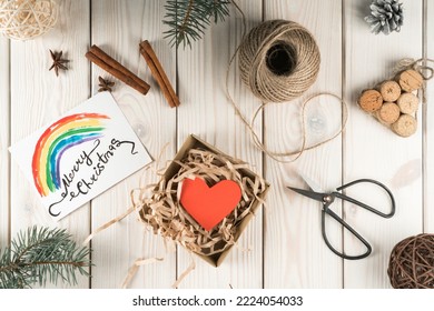 Open Fabric Present Box With Red Heart And Rainbow Label Card Merry Christmas. DIY, Handmade Gifts With Scissors And Wrapping Paper With Fir, Nuts And Cinnamon Decorations On Wooden Background