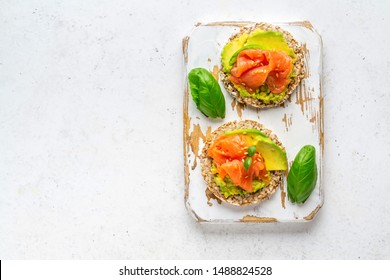 Open face sandwiches on buckwheat crispbread toast with avocado, salmon, basil, flax seeds served on wooden rustic board. Health care, super food concept. White stone background. Top view. Copy space. - Powered by Shutterstock