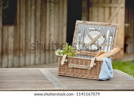 Open fitted wicker picnic hamper with plates, cutlery and French baguettes on a garden table outside a timber cabin with copy space
