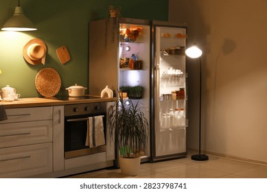 Open fridge full of different fresh products with stove and utensils on counter near green wall in kitchen at night