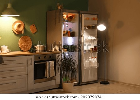 Open fridge full of different fresh products with stove and utensils on counter near green wall in kitchen at night