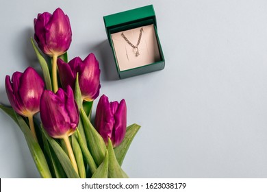 Open Gift Box For Jewelry With A Chain And Pendant Made Of Silver And Fresh Bright Purple Tulips On A Pale Blue Background With Copyspace For Text, Overhead Flat Lay. Can Be Used As A Greeting Ca