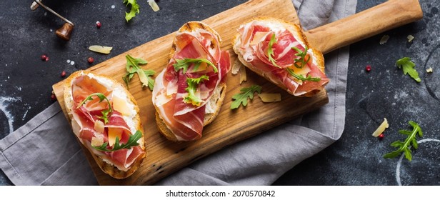 Open ham sandwiches, arugula and hard cheese, served on wooden stand with aglass of red wine on aconcrete old dark background. Rustik style. Top view. - Shutterstock ID 2070570842
