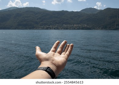 An open hand reaching out towards a serene lake surrounded by lush green mountains under a sunny sky with scattered clouds. - Powered by Shutterstock