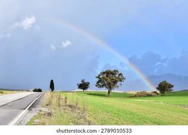 An open landscape shot looking down an asphalt country road stretching alongside green rolling fields with a vibrant rainbow arching over distant mountains under a cloudy sky.