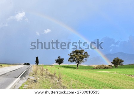 An open landscape shot looking down an asphalt country road stretching alongside green rolling fields with a vibrant rainbow arching over distant mountains under a cloudy sky.
