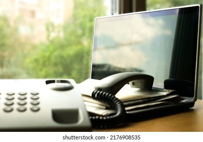 Open Laptop Computer With Stack Of Newspaper And Landline Phone Standing On Office Table On Background Of Window Frame