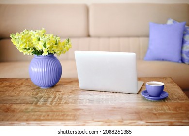 Open laptop, cup of coffee and very peri vase with beautiful yellow flowers on retro wood textured table in minimalistic living room with beige couch in background. Freelance workplace at home