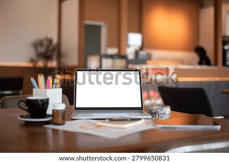 Open laptop displaying empty white screen on a warm wooden desk in a modern cafe, ideal for technology mockup and remote work concepts.