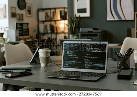 Open laptop displaying programming code on screen sitting on desk in modern office workspace with coffee cup, smartphone, tablet and shelves in background, no people visible