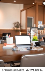 Open laptop with empty white screen mockup on a wooden cafe desk with coffee, pencils, books, and plant, ideal for remote work and freelance concepts.