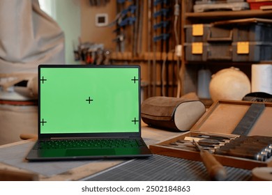 Open laptop with green screen positioned on woodworking desk surrounded by various tools and workshop items including chisel set, mallet, and wooden carvings - Powered by Shutterstock