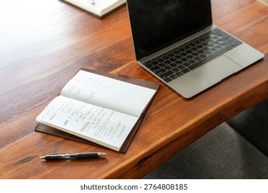 Open laptop and notebook with handwritten notes on a wooden table, accompanied by a pen, suggesting a workspace or study area.