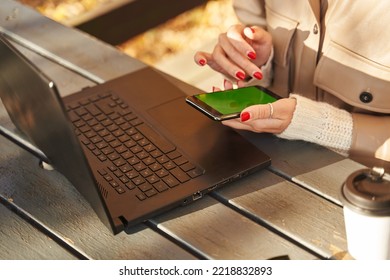 Open Laptop On The Table And A Smartphone With A Green Screen In Female Hands. There Is A Paper Cup With Coffee Next To It