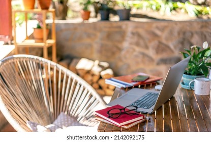 Open laptop on a wooden table, next to glasses on a stack of books on the terrace of a country house on a summer day - Powered by Shutterstock