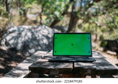 Open Laptop On A Wooden Table In A Camping In The Mountains. A Laptop With A Green Screen Stands On A Camping Table Outdoors Among The Trees Against The Background Of A Car With Travel Things