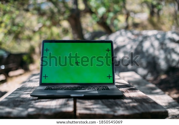 Open laptop on a wooden table in a camping in the\
mountains. A laptop with a green screen stands on a camping table\
outdoors among the trees against the background of a car with\
travel things
