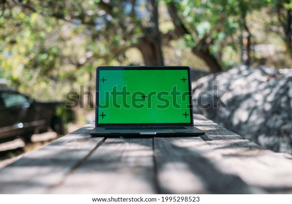 Open laptop on a wooden table in a camping in the\
mountains. A laptop with a green screen stands on a camping table\
outdoors among the trees against the background of a car with\
travel things
