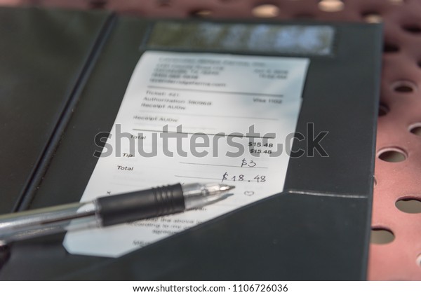 Open leather bill holder with restaurant check\
and pen. Soft focus receipt with hand written total amount and\
tipping on outdoor picnic table, natural light. Paper invoice with\
suggested gratuities