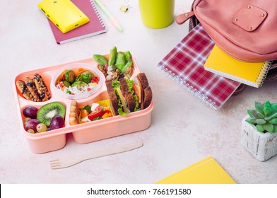 Open Lunch Box With Healthy Lunch With Sandwiches, Vegetable Salad, Fresh Grape And Kiwi, Rice With Grilled Brussels Sprouts And Nuts On The Table Near School Backpack; Selective Focus