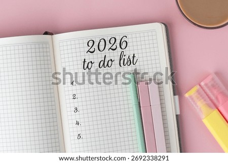 Open notebook with 2026 to do list written on graph paper, surrounded by pastel pens, highlighters, cup of coffee on pink background symbol of planning, organization, and goal setting for the new year
