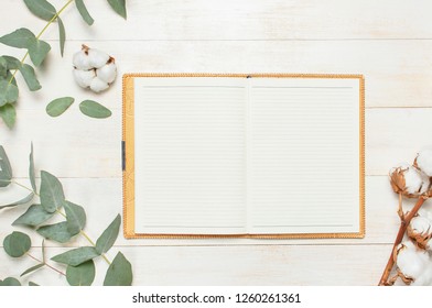 Open notebook with blank pages, pen, eucalyptus twig and cotton flowers on white wooden background top view flat lay. Fashion female blogger working desk. Cotton flowers. Lifestyle gentle background