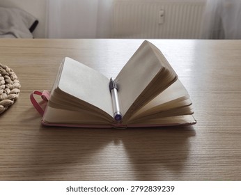 Open notebook with empty pages and a purple pen on a wooden table, accompanied by a woven coaster, illuminated by natural light from a nearby window