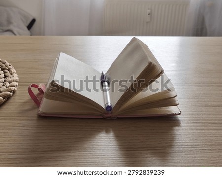 Open notebook with empty pages and a purple pen on a wooden table, accompanied by a woven coaster, illuminated by natural light from a nearby window