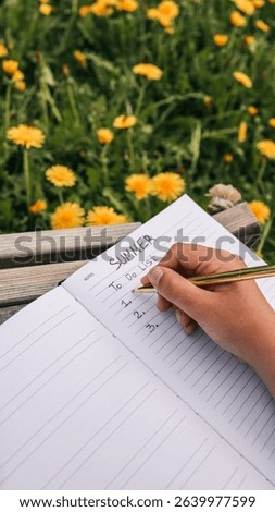Open notebook with "Summer To Do List" written inside, resting on a wooden bench in a field of blooming dandelions, with a straw hat placed nearby in soft afternoon sunlight.