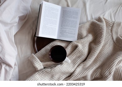 Open Paper Book With Cup Of Coffee On Wooden Tray And Knit Cloth Textile In Bed Close Up Top View. Cozy Home Atmosphere. 